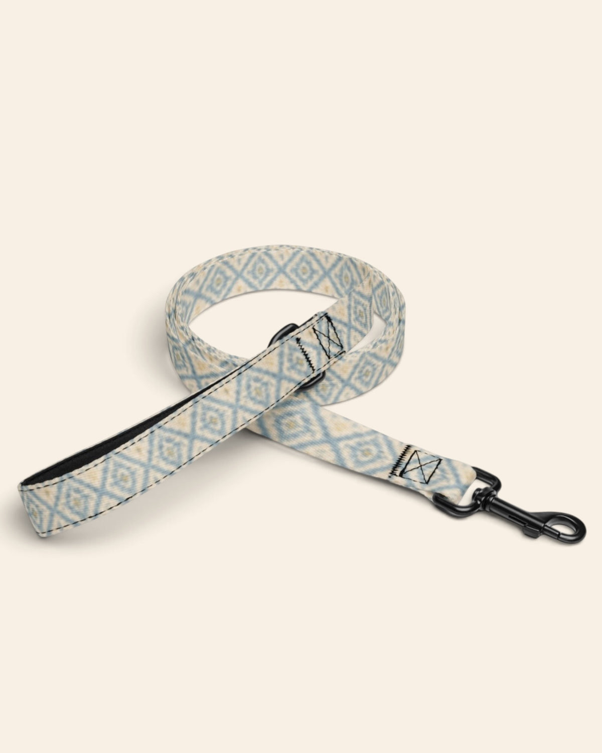The leash coiled on a plain cream background, showing the handle loop, the adjustable slider and the black swivel snap hook. The webbing is cream, printed with dusty blue ikat diamonds picked out with gold and small cross motifs.