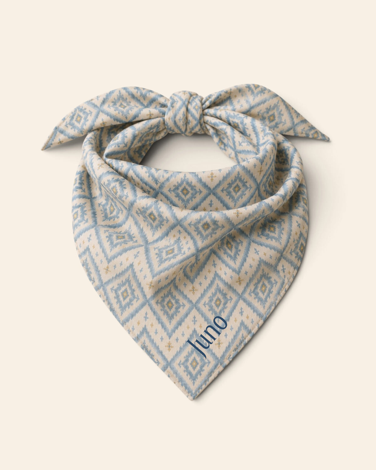 The bandana alone on a plain cream background, knotted so the two tail ends stand up and the triangle hangs below. It is cream, printed with dusty blue ikat diamonds picked out with gold and small cross motifs, with the sample name Juno printed on the lower right panel.