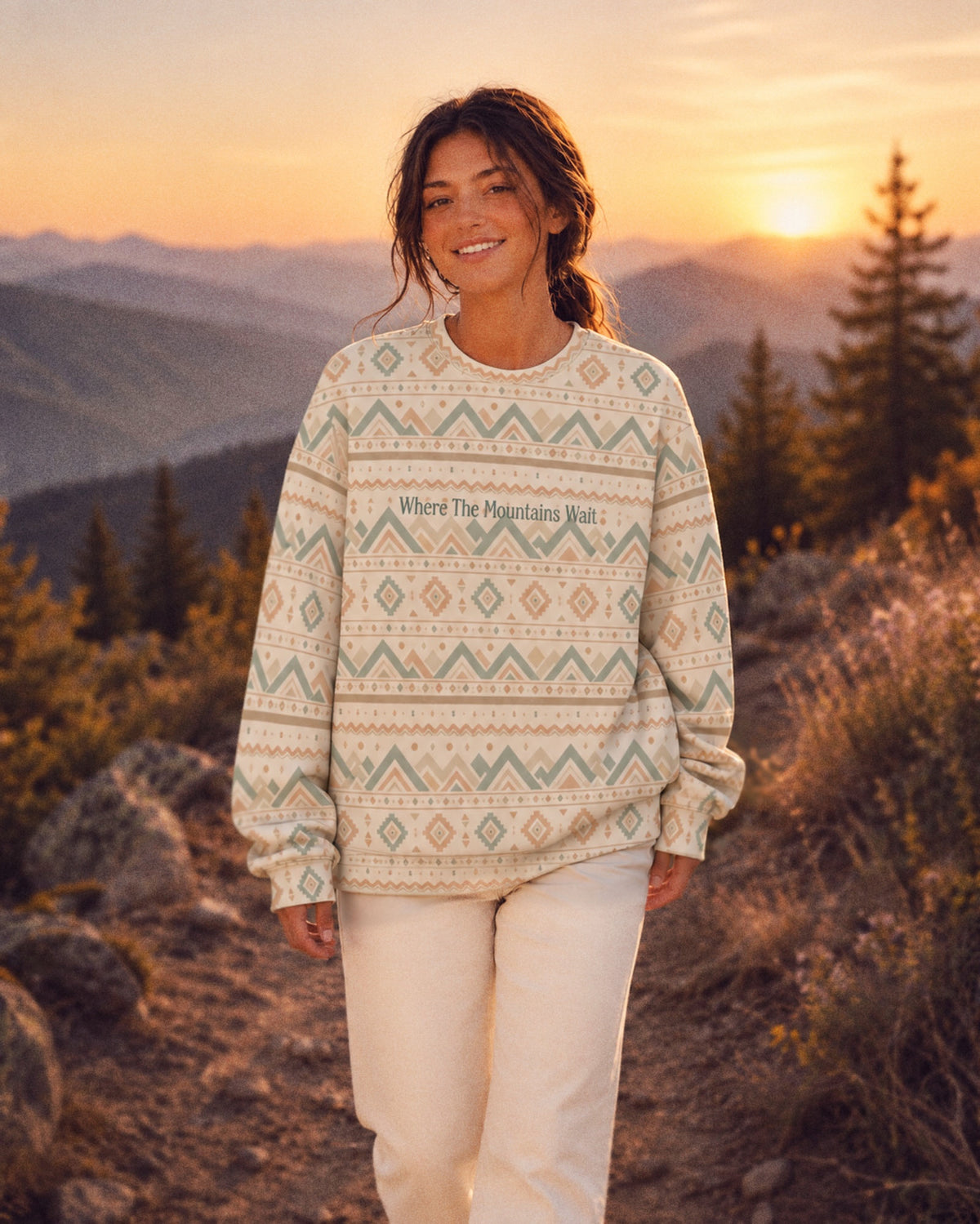 A woman walks along a rocky ridge at sunset, layered mountain ranges and conifers behind her wearing the oversized crewneck sweatshirt. It is cream, banded with sage and gold chevrons, diamonds and small geometric motifs, with the words Where The Mountains Wait printed in a small script across the chest.