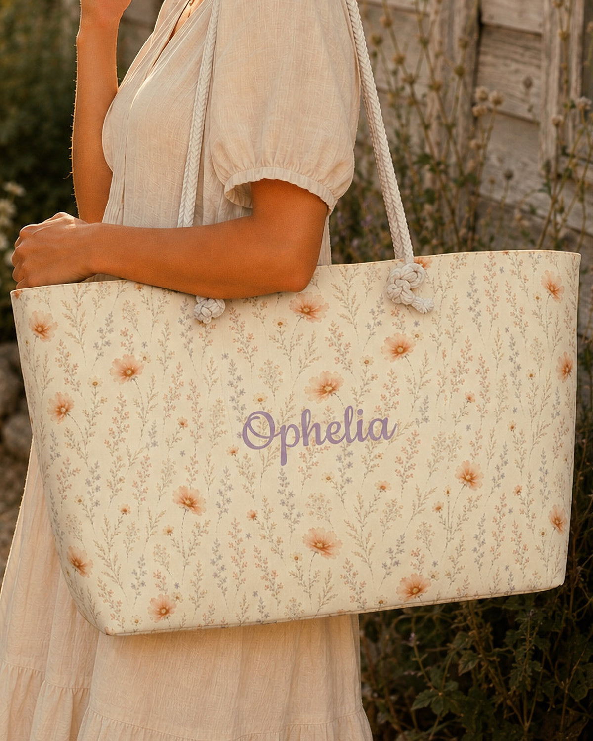 A woman carries the weekender tote on her forearm, wearing a cream linen dress, beside a weathered timber post in warm afternoon light. The bag is cream, scattered with small apricot daisies among sage stems, with the sample name Ophelia in a flowing script across the side panel and thick cream rope handles knotted through the top corners.