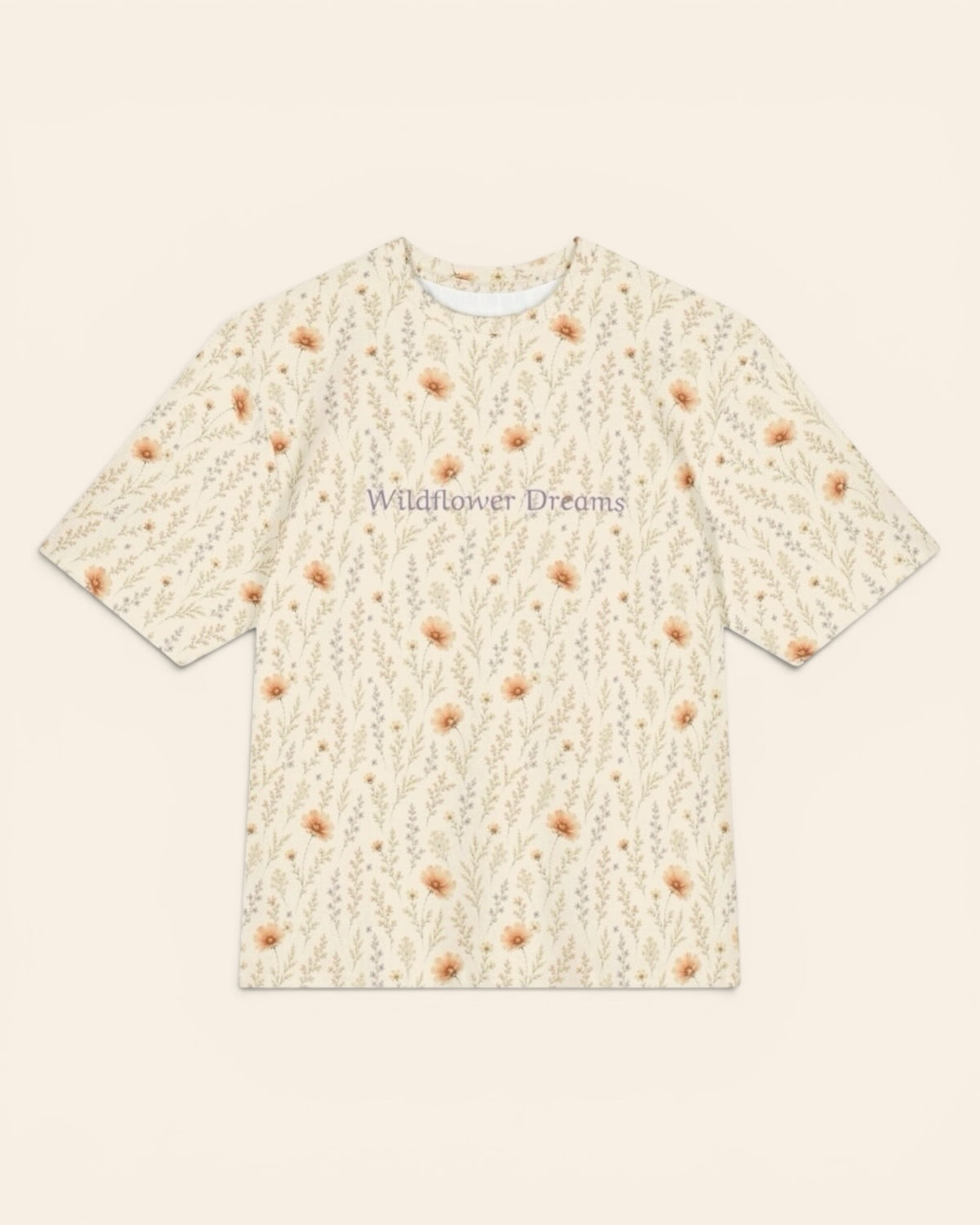 The oversized tee shown flat from the front on a plain cream background, short sleeves squared out from a relaxed body. The all-over print is warm cream, printed with apricot cosmos blooms among fine sage stems, with the words Wildflower Dreams in a small script across the chest.