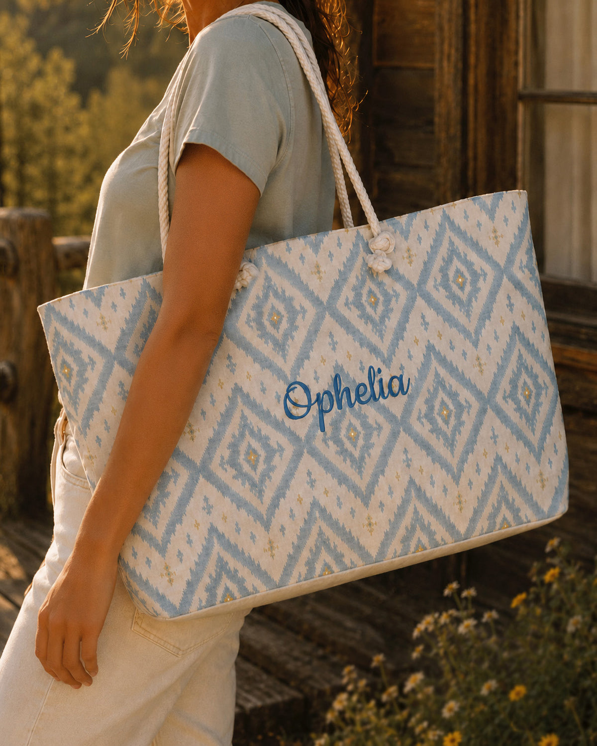 A woman carries the weekender tote at her side, wearing a sage tee, in dry grass beside a weathered barn. The bag is cream, printed with a lattice of dusty blue ikat diamonds, with the sample name Ophelia in a flowing script across the side panel and thick cream rope handles knotted through the top corners.