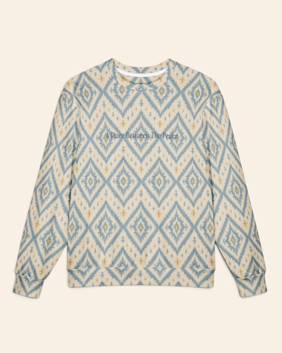 The oversized crewneck sweatshirt shown flat from the front on a plain cream background, sleeves out, cuffs and waistband ribbed. The all-over print is cream, printed with large dusty blue ikat diamonds outlined in gold with small cross motifs, with the words A Place Between The Peaks in a small script across the chest.