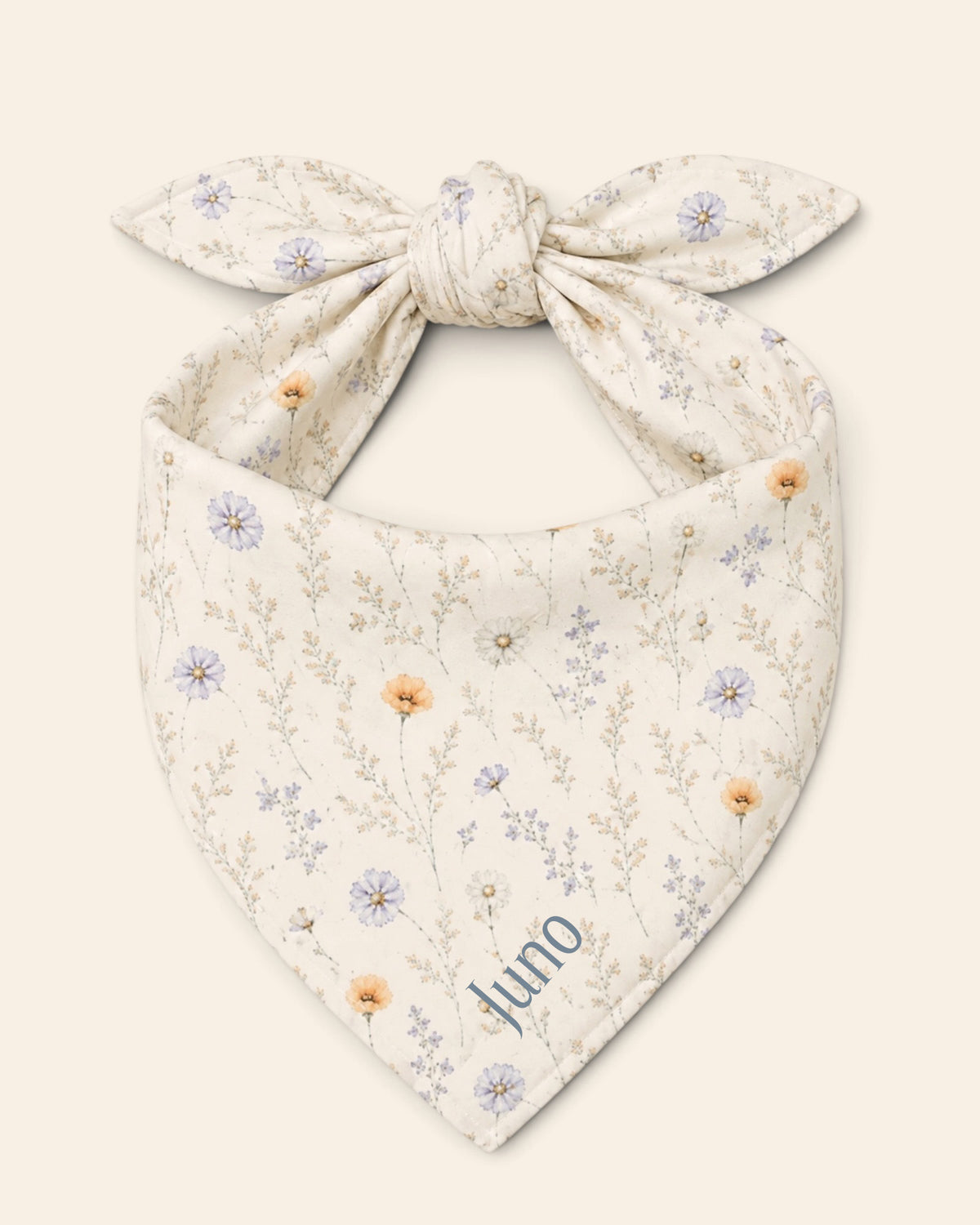 The bandana alone on a plain cream background, knotted so the two tail ends stand up and the triangle hangs below. It is white, printed with small apricot poppies and lilac and periwinkle wildflowers on fine stems, with the sample name Juno printed on the lower right panel.