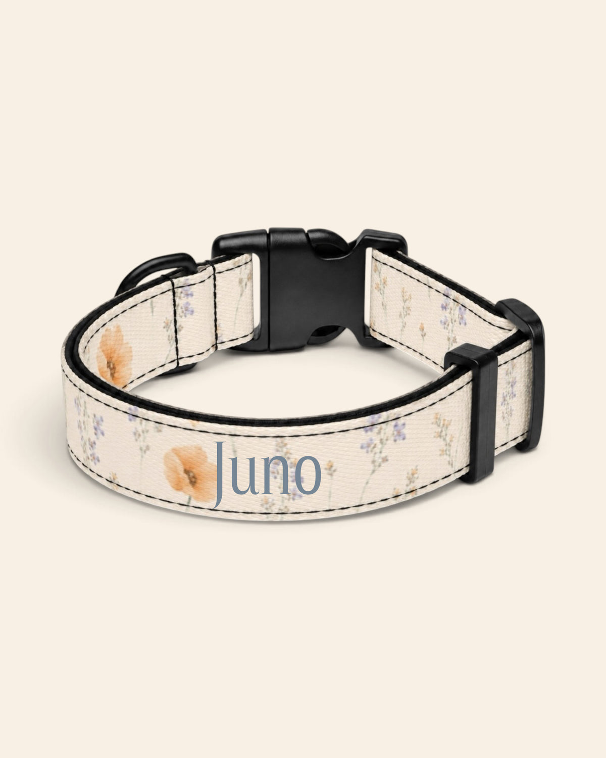 The collar alone on a plain cream background, lying in a loose loop that shows the printed webbing, a black side-release buckle and a black slider. The strap is white, printed with small apricot poppies and lilac and periwinkle wildflowers on fine stems, with the sample name Juno printed along it.