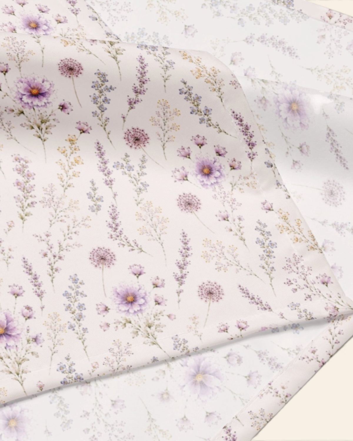 A close crop of the bandana fabric with one corner folded back over a soft cream ground, showing the print at full scale - white, printed with lavender and mauve blooms on slender stems.
