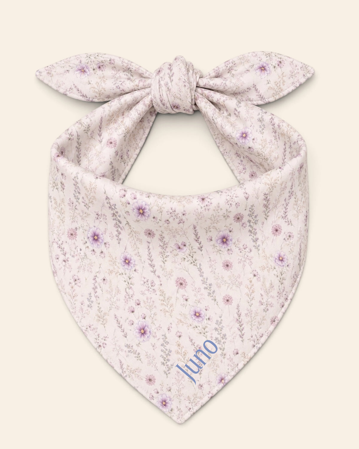 The bandana alone on a plain cream background, knotted so the two tail ends stand up and the triangle hangs below. It is white, printed with lavender and mauve blooms on slender stems, with the sample name Juno printed on the lower right panel.