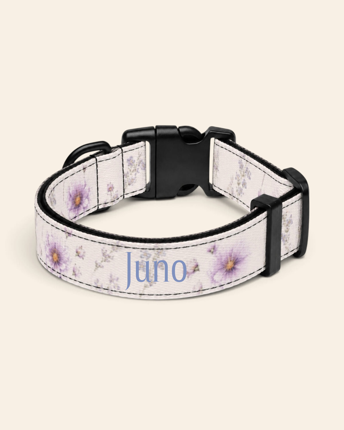 The collar alone on a plain cream background, lying in a loose loop that shows the printed webbing, a black side-release buckle and a black slider. The strap is white, printed with lavender and mauve blooms on slender stems, with the sample name Juno printed along it.