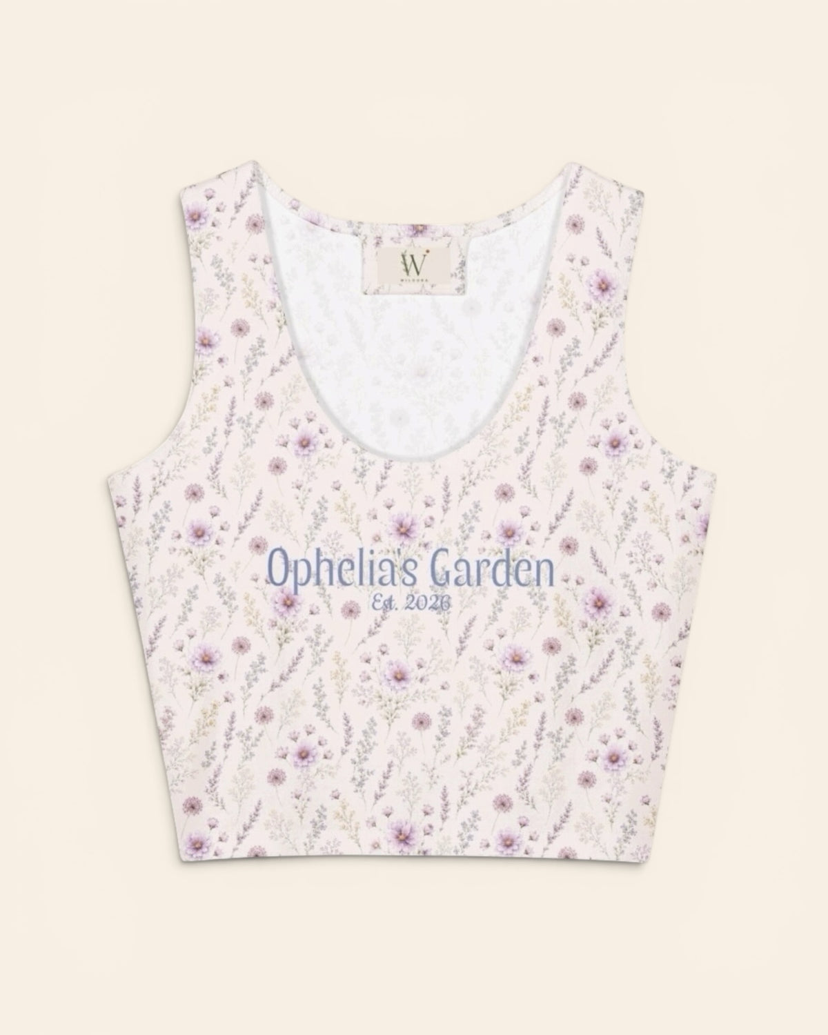 The crop tank shown from the front on a plain cream background, a scoop neck lined in white with a small woven W label at the back collar. The print is white, printed with lavender and mauve wildflowers on slender stems, and the two-line caption “Ophelia’s Garden” above “Est. 2026” is printed in a small script across the chest.