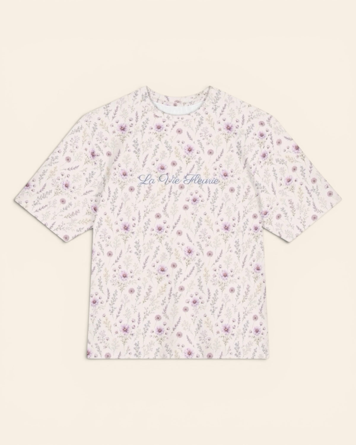 The oversized tee shown flat from the front on a plain cream background, short sleeves squared out from a relaxed body. The all-over print is white, printed with lavender and mauve wildflowers on slender stems, with the words La Vie Fleurie in a small script across the chest.