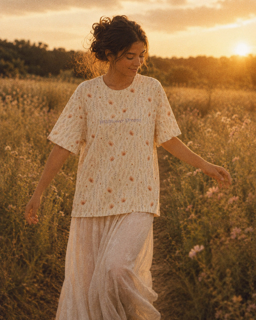 A woman walks through a meadow of dry grass at sunset wearing the oversized tee, cut long and loose with a dropped shoulder. It is warm cream, printed with apricot cosmos blooms among fine sage stems, with the words Wildflower Dreams printed in a small script across the chest.