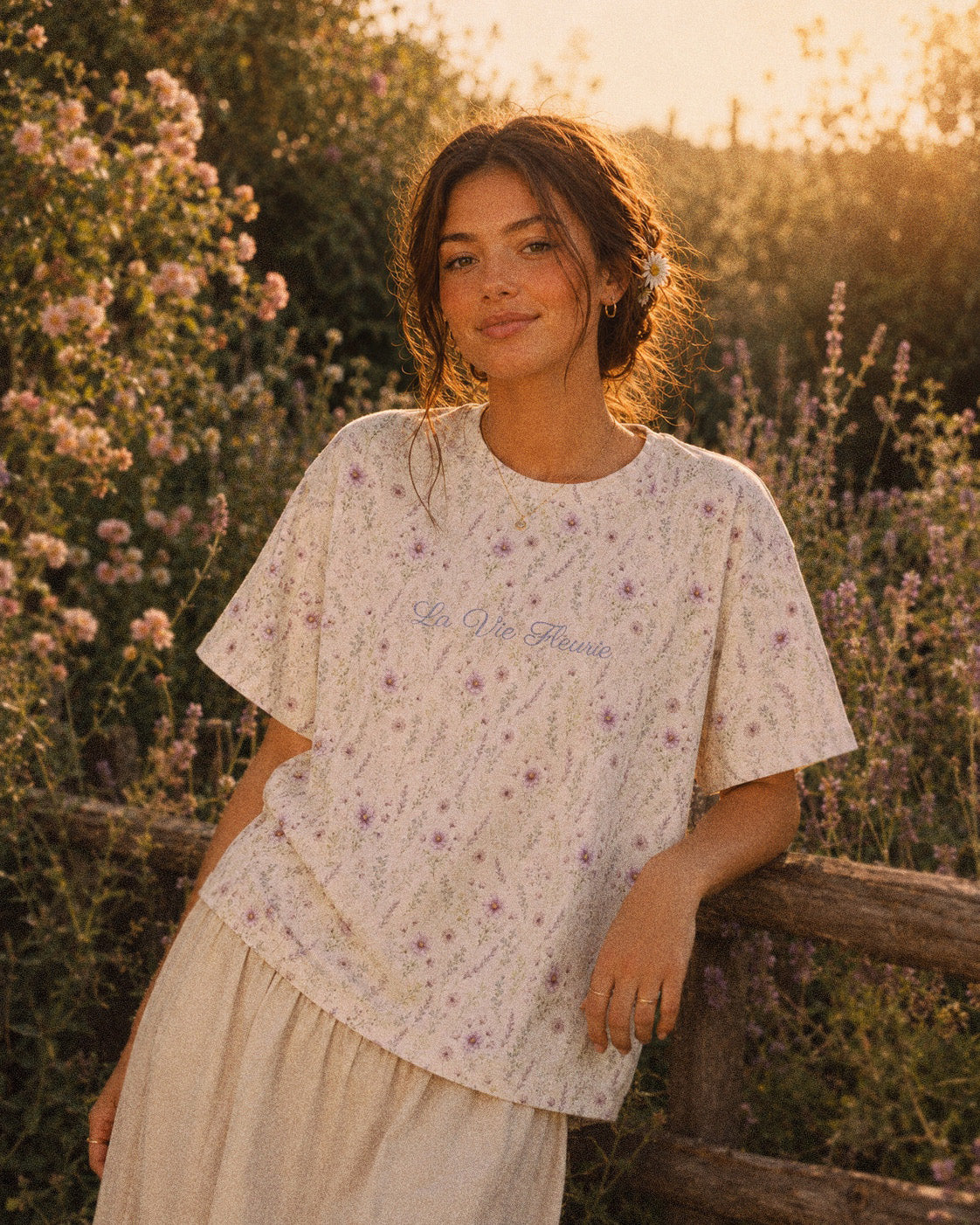 A woman walks beside blossoming shrubs at golden hour wearing the oversized tee, cut long and loose with a dropped shoulder. It is white, printed with lavender and mauve wildflowers on slender stems, with the words La Vie Fleurie printed in a small script across the chest.