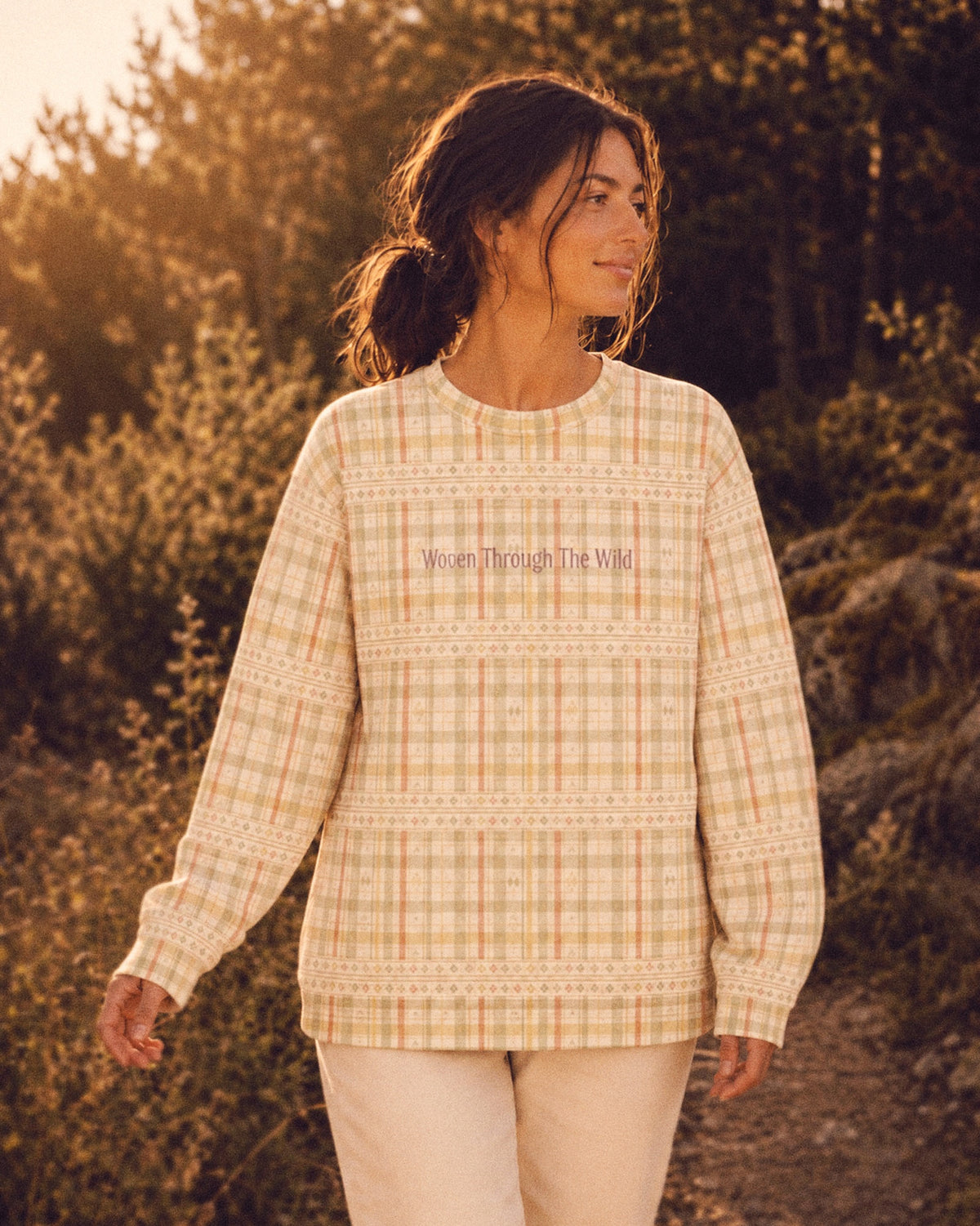 A woman walks among sunlit shrubs and dry grasses at golden hour wearing the oversized crewneck sweatshirt. It is a sage, gold and apricot plaid on cream, banded with rows of small woven motifs, with the words Woven Through The Wild printed in a small script across the chest.
