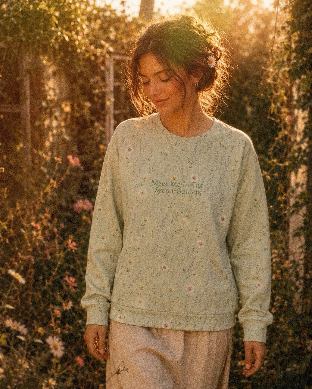 A woman walks beneath a flowering arbour in golden light wearing the oversized crewneck sweatshirt. It is sage green, printed with white and pink daisies and fine wildflower stems, with the words Meet Me In The Secret Garden printed in a small script across the chest.