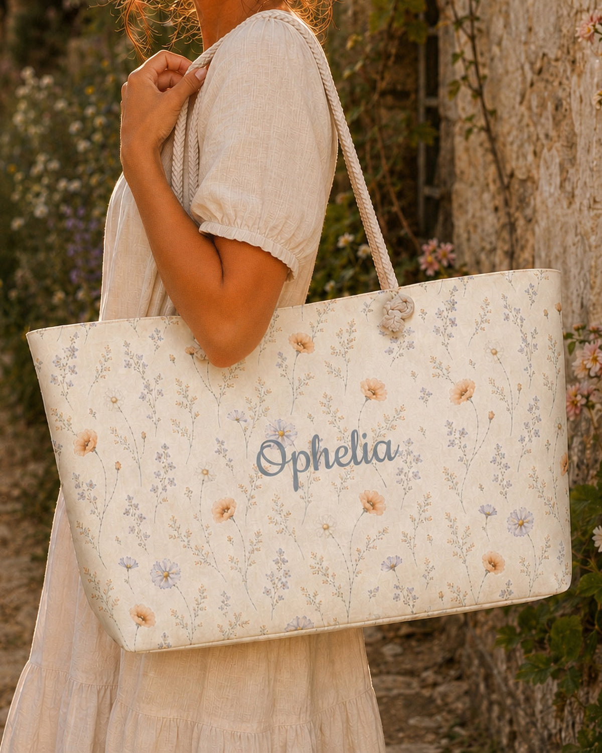 A woman carries the weekender tote on her shoulder, wearing a cream linen dress, against rustic weathered boards in golden light. The bag is white, scattered with apricot and lilac wildflowers on green stems, with the sample name Ophelia in a flowing script across the side panel and thick cream rope handles knotted through the top corners.