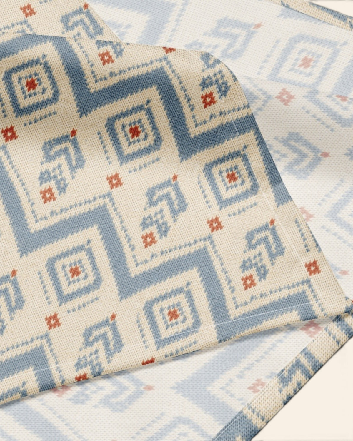 A close crop of the bandana fabric with one corner folded back over a soft cream ground, showing the print at full scale - cream, printed with dusty blue zigzag chevrons and stepped diamond motifs with small rust accents.