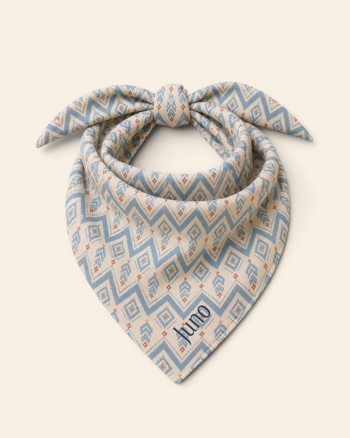 The bandana alone on a plain cream background, knotted so the two tail ends stand up and the triangle hangs below. It is cream, printed with dusty blue zigzag chevrons and stepped diamond motifs with small rust accents, with the sample name Juno printed on the lower right panel.