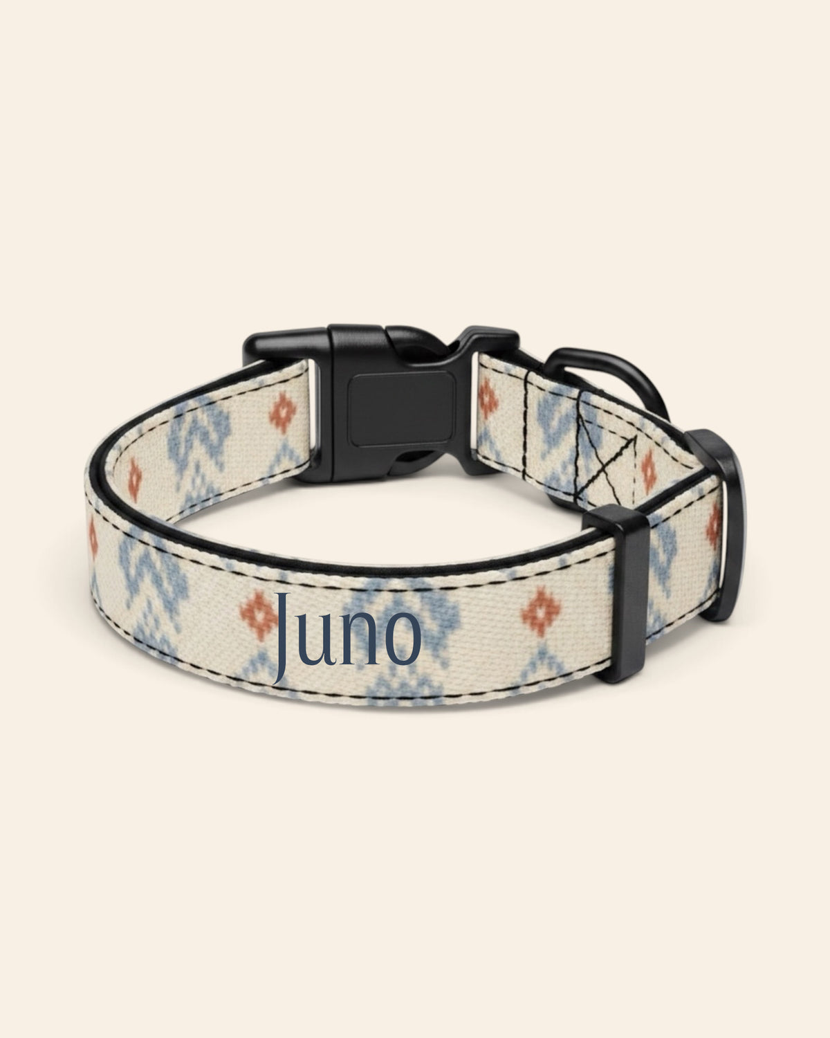 The collar alone on a plain cream background, lying in a loose loop that shows the printed webbing, a black side-release buckle and a black slider. The strap is cream, printed with dusty blue zigzag chevrons and stepped diamond motifs with small rust accents, with the sample name Juno printed along it.
