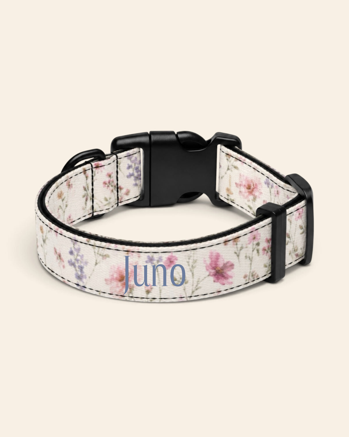 The collar alone on a plain cream background, lying in a loose loop that shows the printed webbing, a black side-release buckle and a black slider. The strap is white, densely printed with pink, lilac and periwinkle wildflowers, with the sample name Juno printed along it.