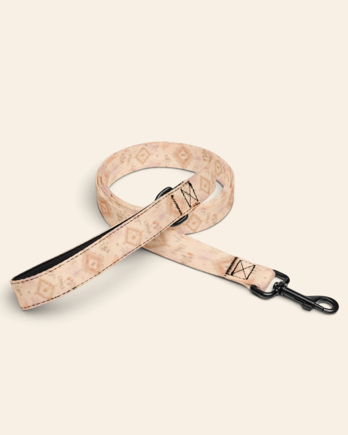 The leash coiled on a plain cream background, showing the handle loop, the adjustable slider and the black swivel snap hook. The webbing is an apricot and peach wash set with cream diamond medallions and fine sprigs.