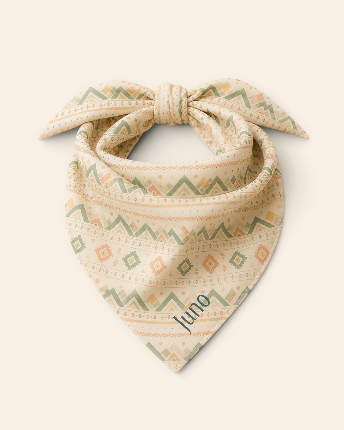 The bandana alone on a plain cream background, knotted so the two tail ends stand up and the triangle hangs below. It is cream, banded with sage and apricot chevrons and diamond geometric motifs, with the sample name Juno printed on the lower right panel.