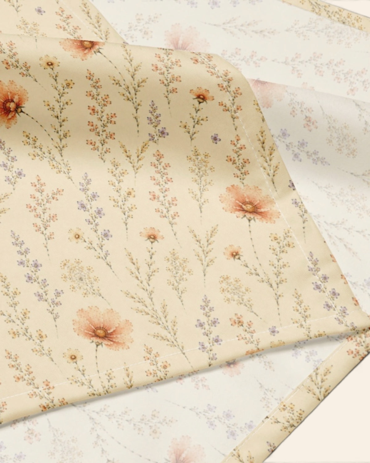 A close crop of the bandana fabric with one corner folded back over a soft cream ground, showing the print at full scale - soft butter-cream, printed with small apricot and coral blooms among fine sage stems.