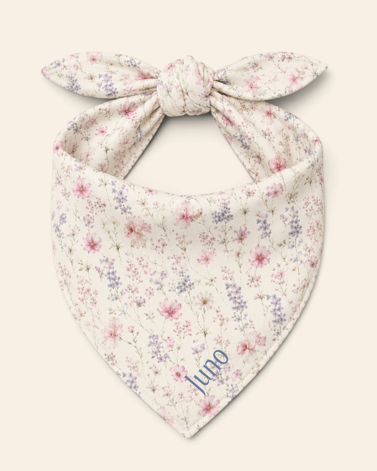 The bandana alone on a plain cream background, knotted so the two tail ends stand up and the triangle hangs below. It is white, densely printed with pink, lilac and periwinkle wildflowers, with the sample name Juno printed on the lower right panel.