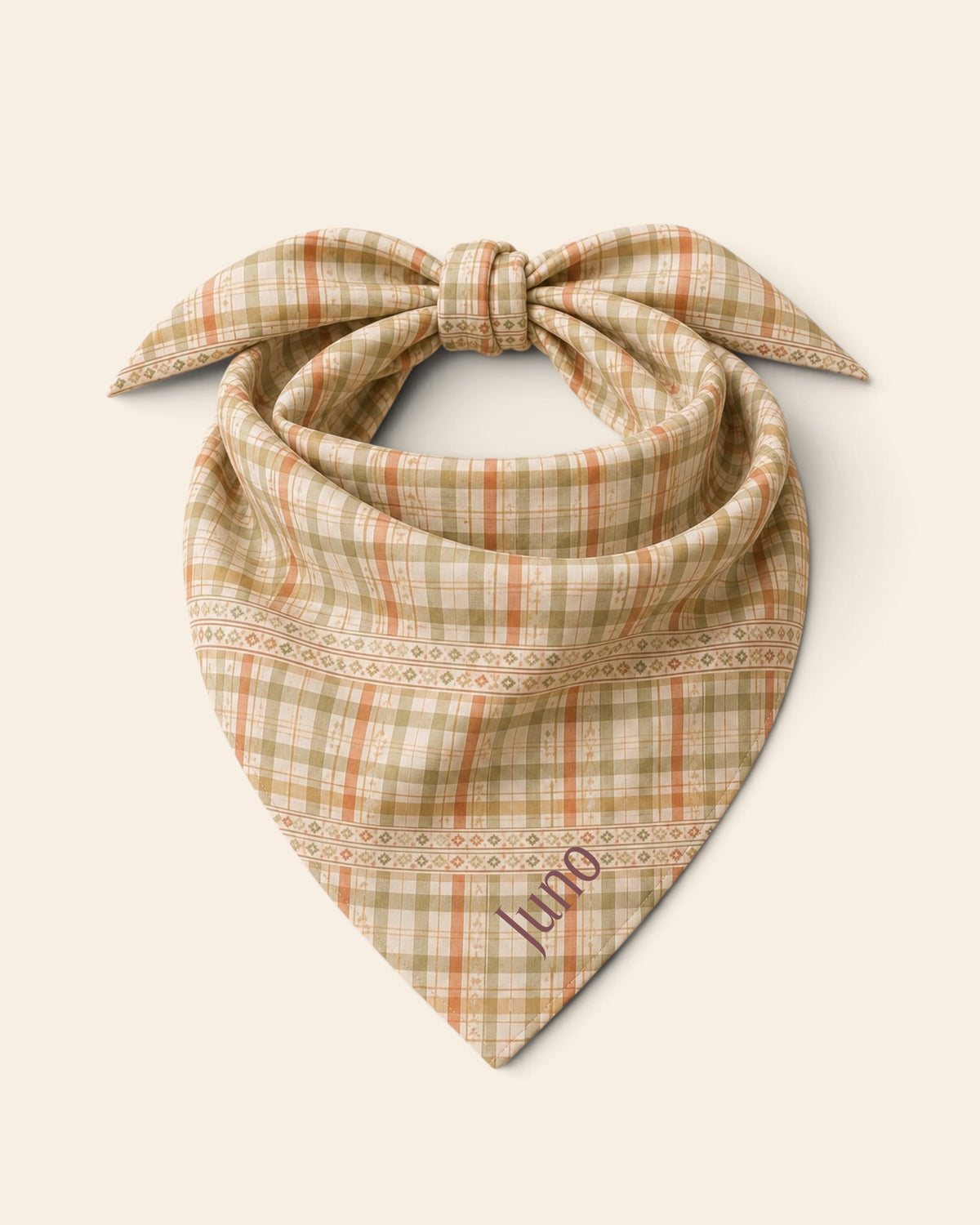 The bandana alone on a plain cream background, knotted so the two tail ends stand up and the triangle hangs below. It is a sage, gold and apricot plaid on cream, banded with rows of small motifs, with the sample name Juno printed on the lower right panel.