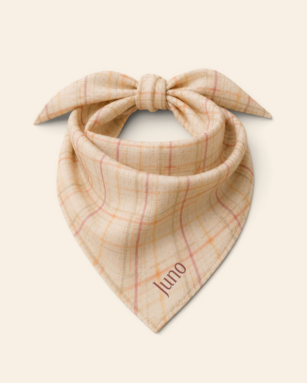 The bandana alone on a plain cream background, knotted so the two tail ends stand up and the triangle hangs below. It is a cream plaid crossed by apricot, rose and sage lines, with the sample name Juno printed on the lower right panel.