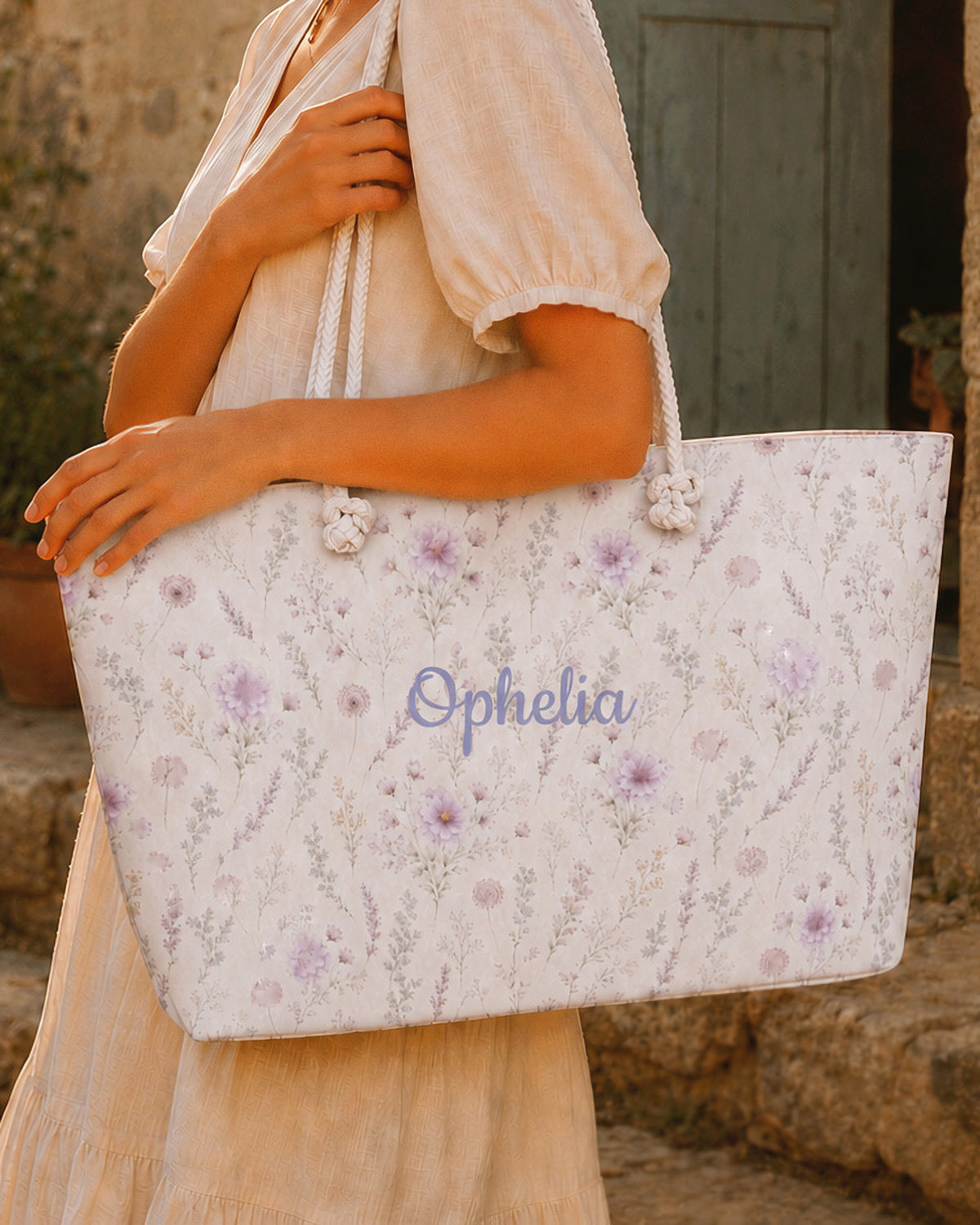 A woman carries the weekender tote on her forearm, wearing a cream linen dress, in front of a weathered blue-grey wooden door and stone wall. The bag is white, printed with lavender and mauve blooms on fine trailing stems, with the sample name Ophelia in a flowing script across the side panel and thick cream rope handles knotted through the top corners.