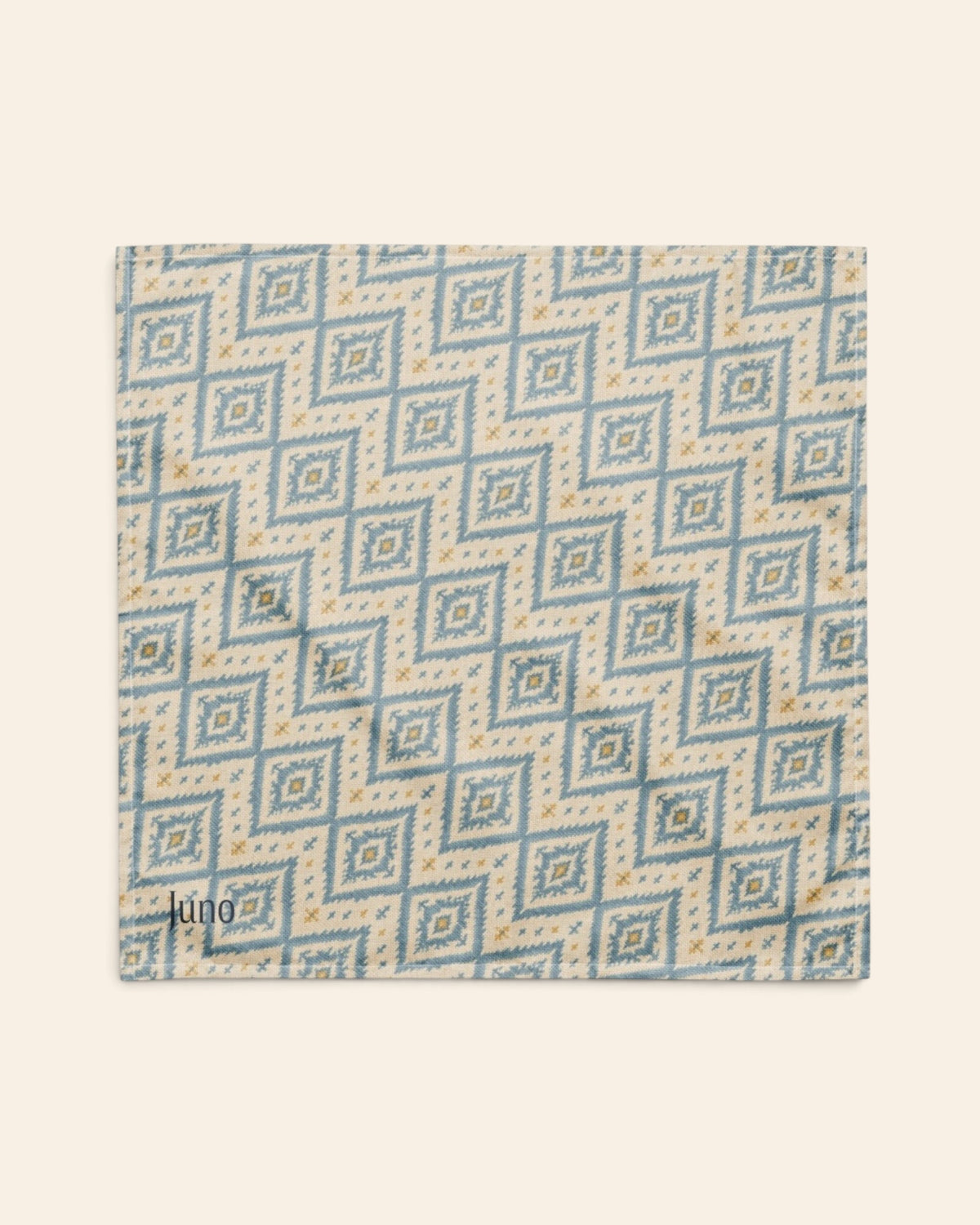 The bandana laid flat as a full square on a cream background, the print running on the diagonal. It is cream, printed with dusty blue ikat diamonds picked out with gold and small cross motifs, with the sample name Juno printed in the lower left corner.