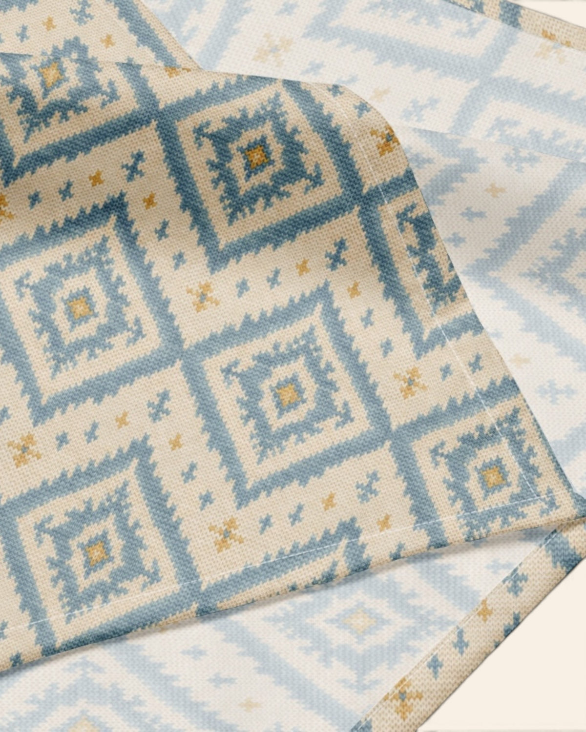 A close crop of the bandana fabric with one corner folded back over a soft cream ground, showing the print at full scale - cream, printed with dusty blue ikat diamonds picked out with gold and small cross motifs.