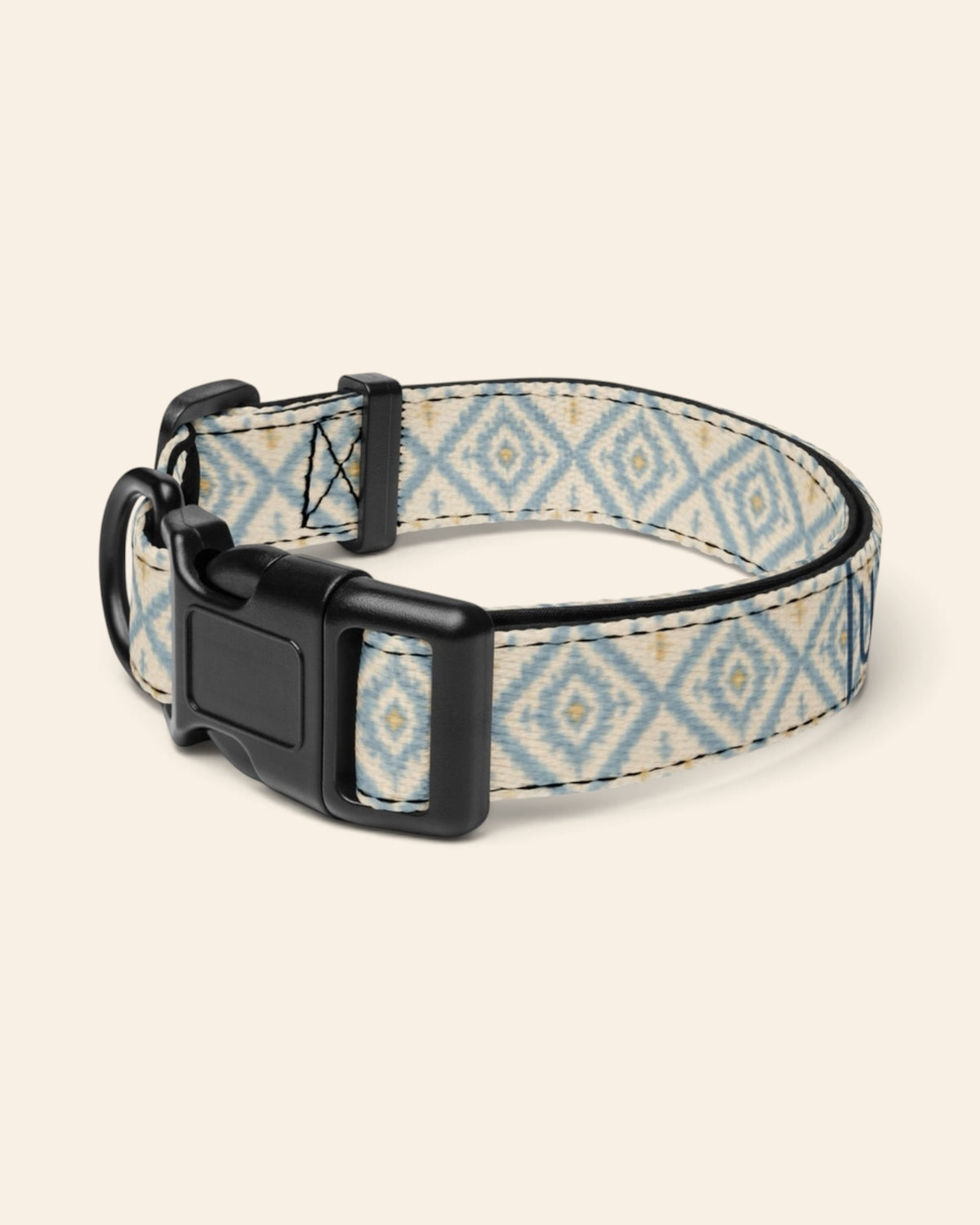 The collar alone on a plain cream background with no personalisation, stood in a ring and angled to show the black side-release buckle face, the D-ring and the tri-glide slider. The webbing is cream, printed with dusty blue ikat diamonds picked out with gold and small cross motifs.