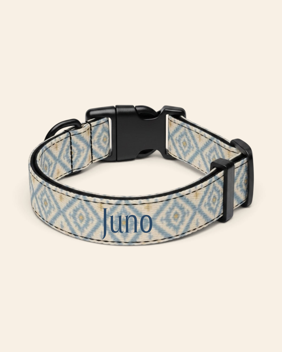 The collar alone on a plain cream background, lying in a loose loop that shows the printed webbing, a black side-release buckle and a black slider. The strap is cream, printed with dusty blue ikat diamonds picked out with gold and small cross motifs, with the sample name Juno printed along it.