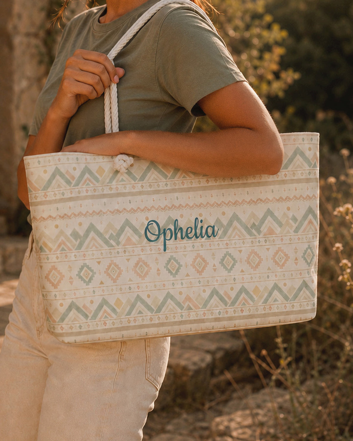 A woman carries the weekender tote at her side, wearing a sage tee, with summer greenery behind. The bag is cream, banded with sage and gold chevrons and diamond geometric motifs, with the sample name Ophelia in a flowing script across the side panel and thick cream rope handles knotted through the top corners.