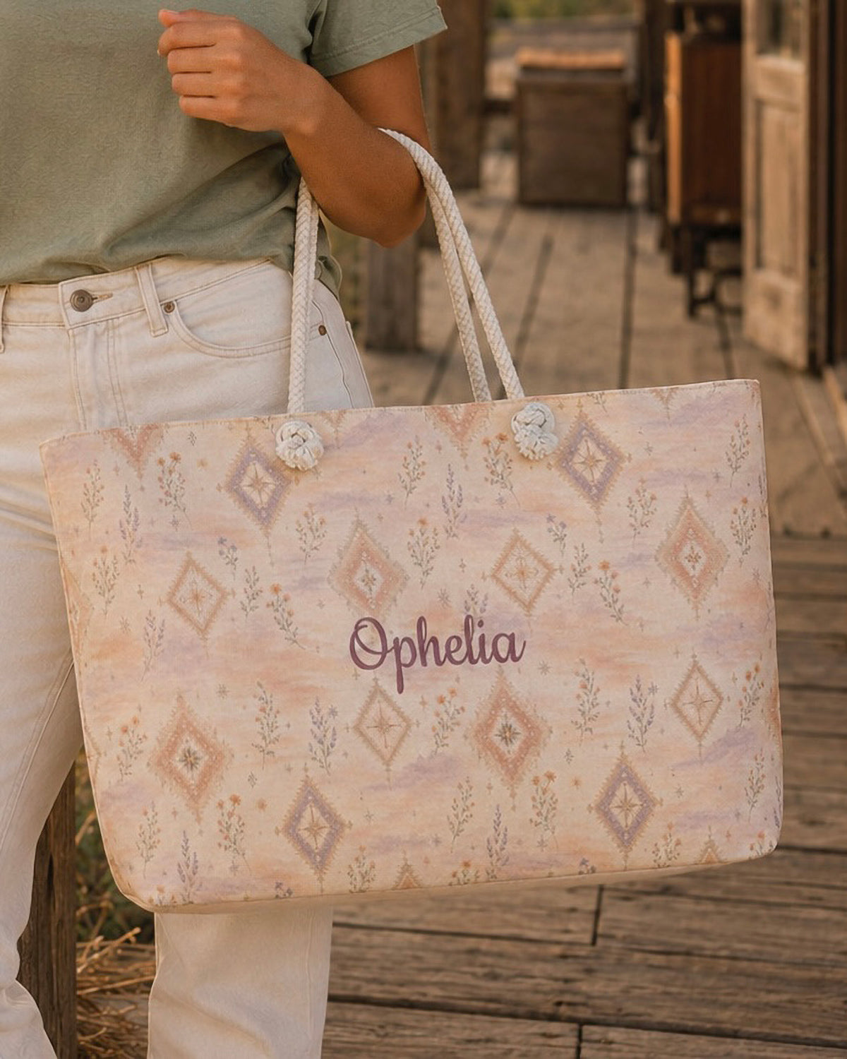 A woman carries the weekender tote at her side, wearing a sage tee and cream trousers, standing on a wooden boardwalk. The bag is a peach and blush wash set with cream diamond medallions and small sprigs, with the sample name Ophelia in a flowing script across the side panel and thick cream rope handles knotted through the top corners.