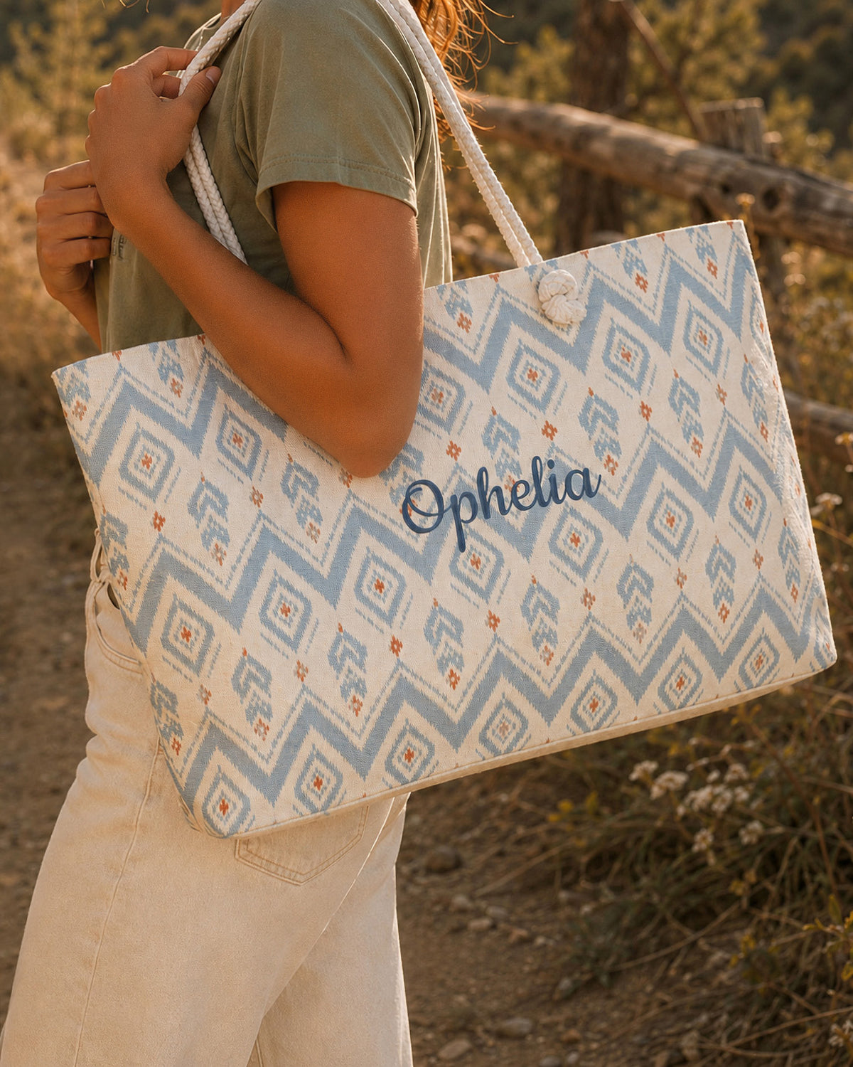 A woman carries the weekender tote at her side, wearing a sage tee, in dry grass and low scrub. The bag is cream, printed with dusty blue zigzag bands and apricot diamond motifs, with the sample name Ophelia in a flowing script across the side panel and thick cream rope handles knotted through the top corners.