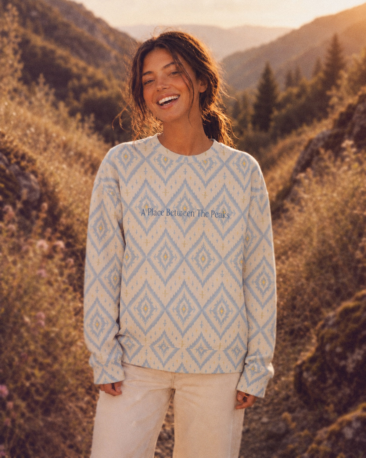 A woman walks across a sunlit grassy mountain slope, conifers and hillsides behind her wearing the oversized crewneck sweatshirt. It is cream, printed with large dusty blue ikat diamonds outlined in gold with small cross motifs, with the words A Place Between The Peaks printed in a small script across the chest.