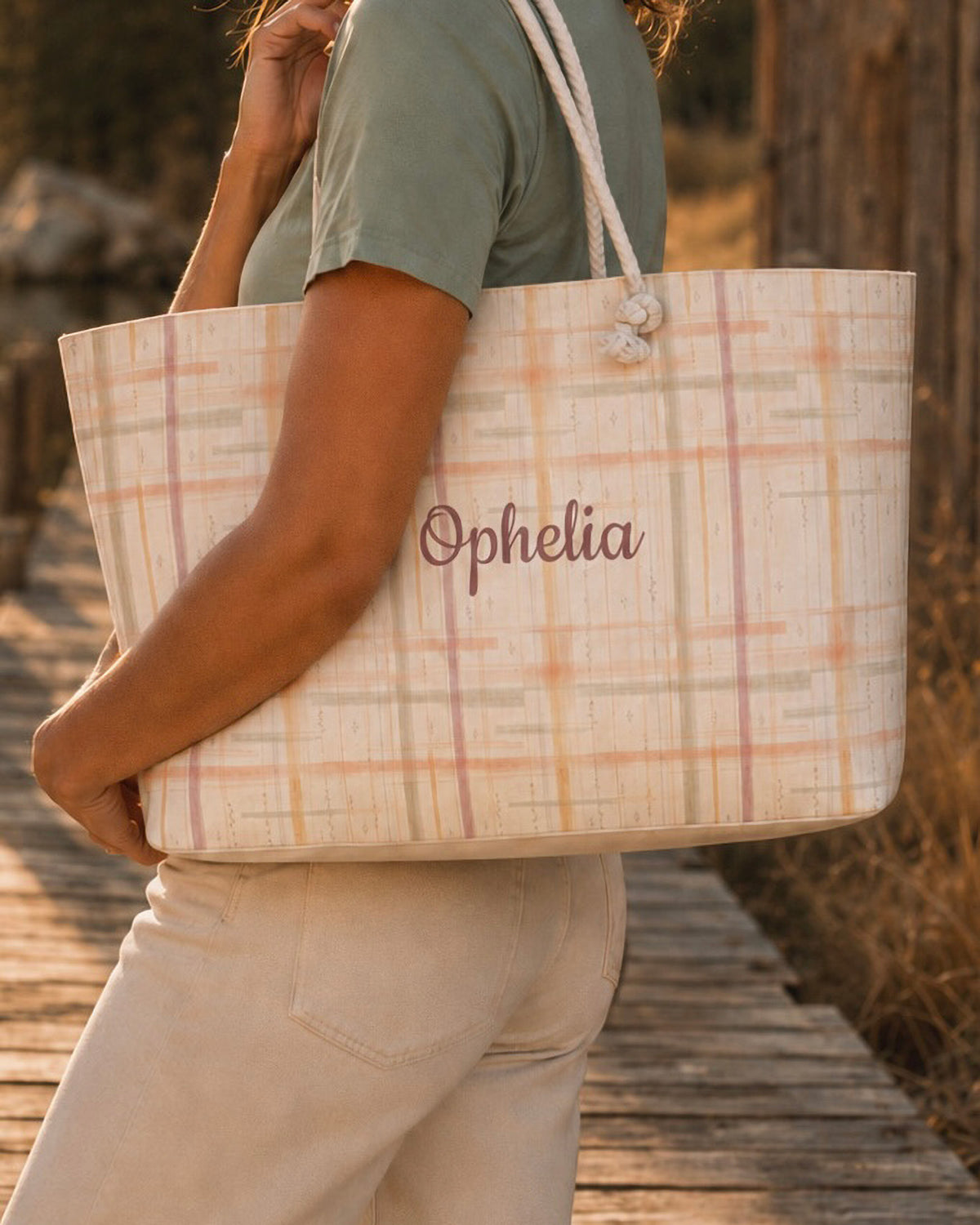 A woman carries the weekender tote at her side, wearing a sage tee and cream trousers, on a wooden boardwalk. The bag is a cream plaid crossed by apricot and coral lines, with the sample name Ophelia in a flowing script across the side panel and thick cream rope handles knotted through the top corners.