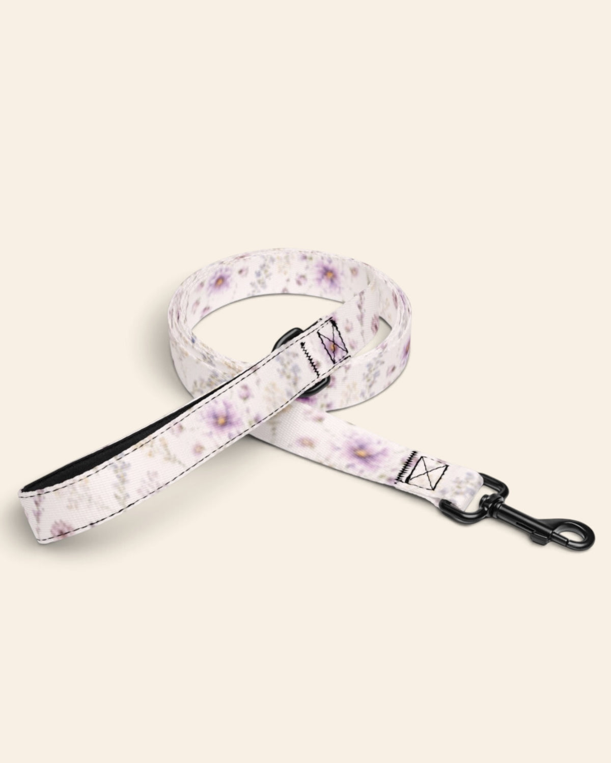 The leash coiled on a plain cream background, showing the handle loop, the adjustable slider and the black swivel snap hook. The webbing is white, printed with lavender and mauve blooms on slender stems.