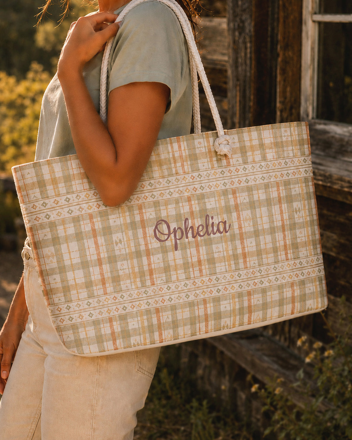 A woman carries the weekender tote at her side, wearing a sage tee, in dry grass beside a weathered barn. The bag is a sage, gold and apricot plaid banded with rows of small dotted motifs, with the sample name Ophelia in a flowing script across the side panel and thick cream rope handles knotted through the top corners.