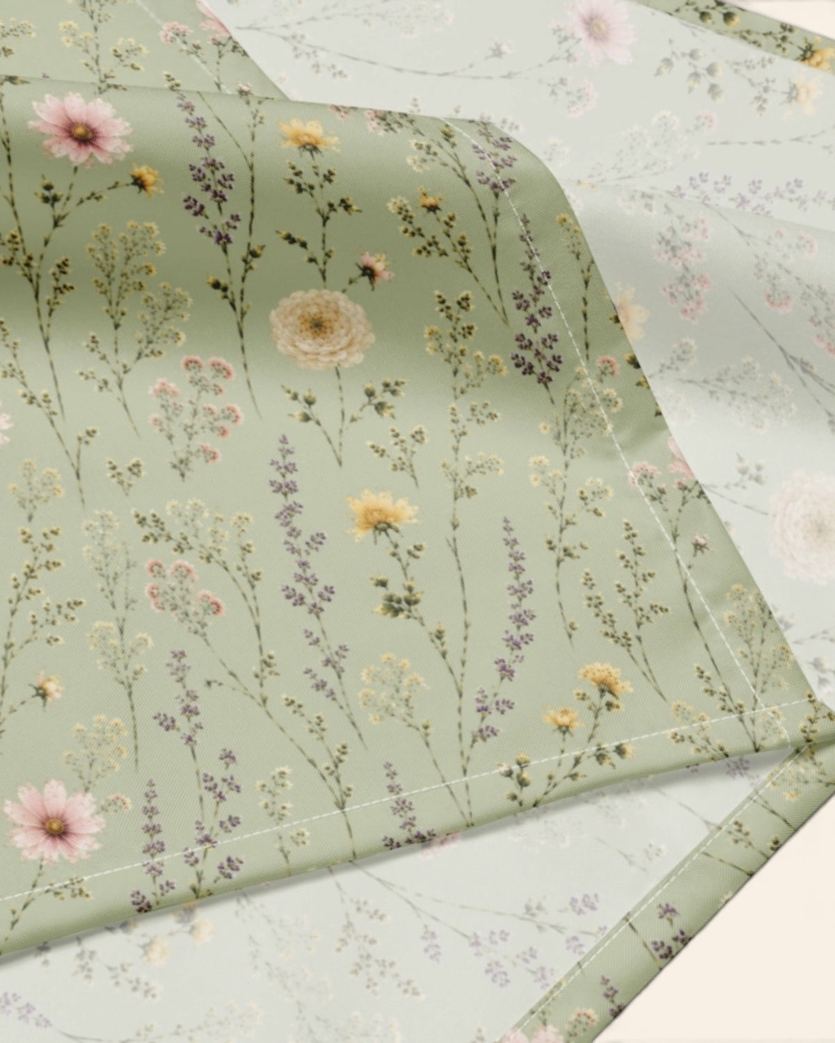 A close crop of the bandana fabric with one corner folded back over a soft cream ground, showing the print at full scale - sage green, printed with small white, pink and yellow blooms on fine stems.