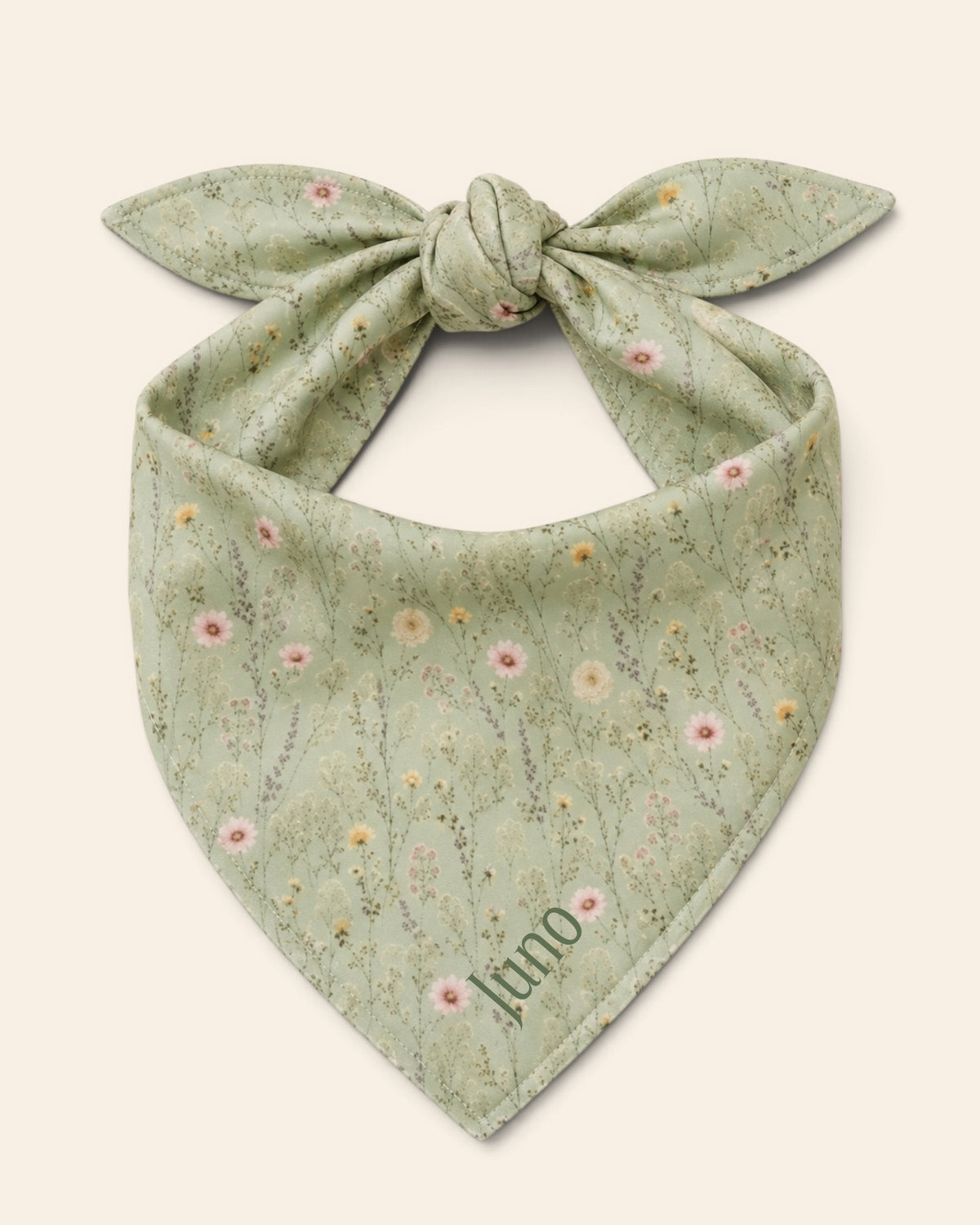 The bandana alone on a plain cream background, knotted so the two tail ends stand up and the triangle hangs below. It is sage green, printed with small white, pink and yellow blooms on fine stems, with the sample name Juno printed on the lower right panel.