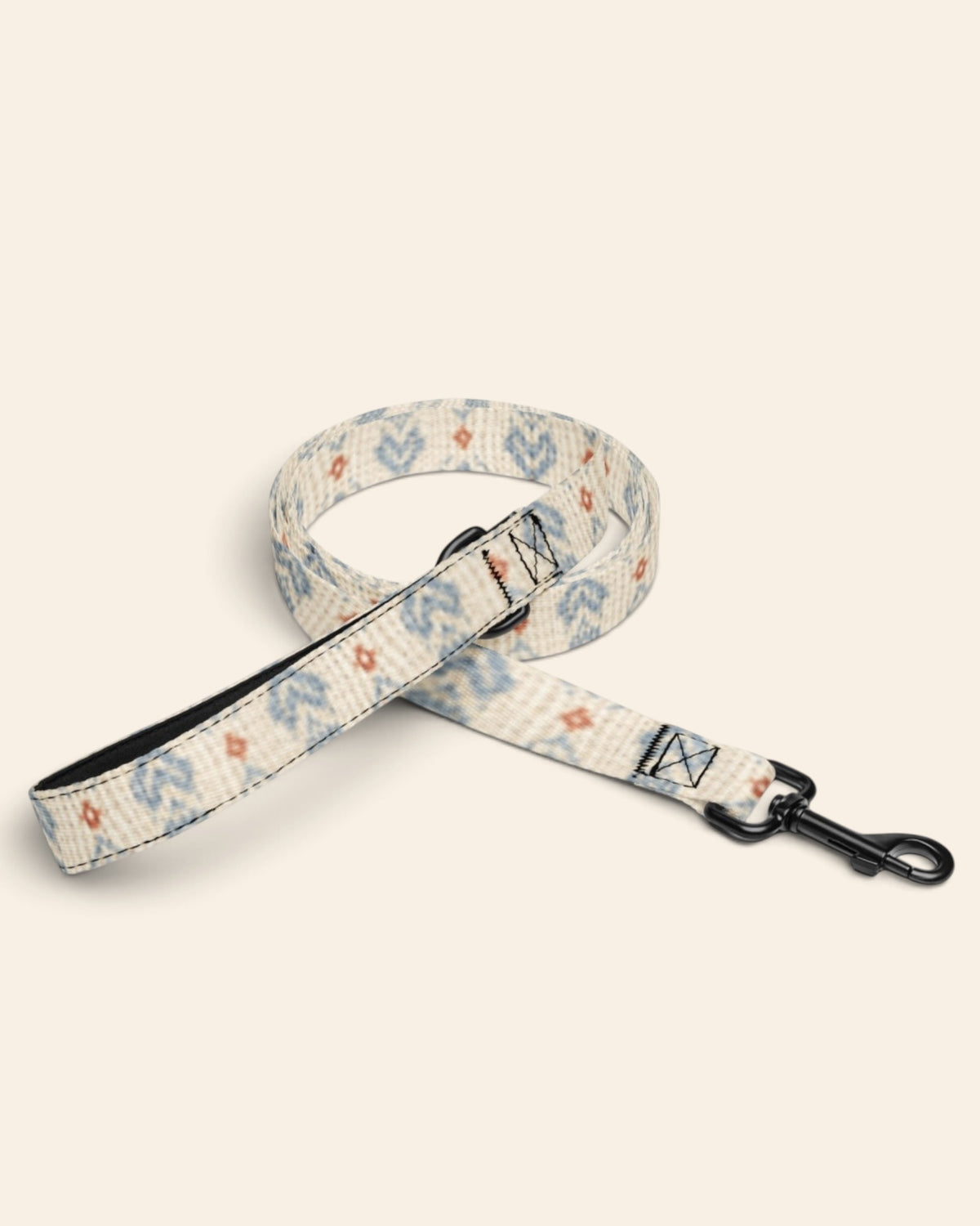 The leash coiled on a plain cream background, showing the handle loop, the adjustable slider and the black swivel snap hook. The webbing is cream, printed with dusty blue zigzag chevrons and stepped diamond motifs with small rust accents.