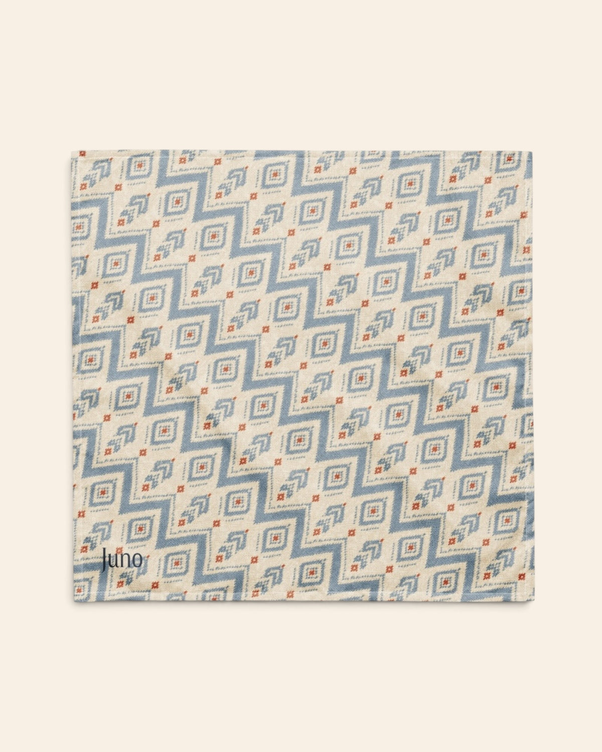 The bandana laid flat as a full square on a cream background, the print running on the diagonal. It is cream, printed with dusty blue zigzag chevrons and stepped diamond motifs with small rust accents, with the sample name Juno printed in the lower left corner.