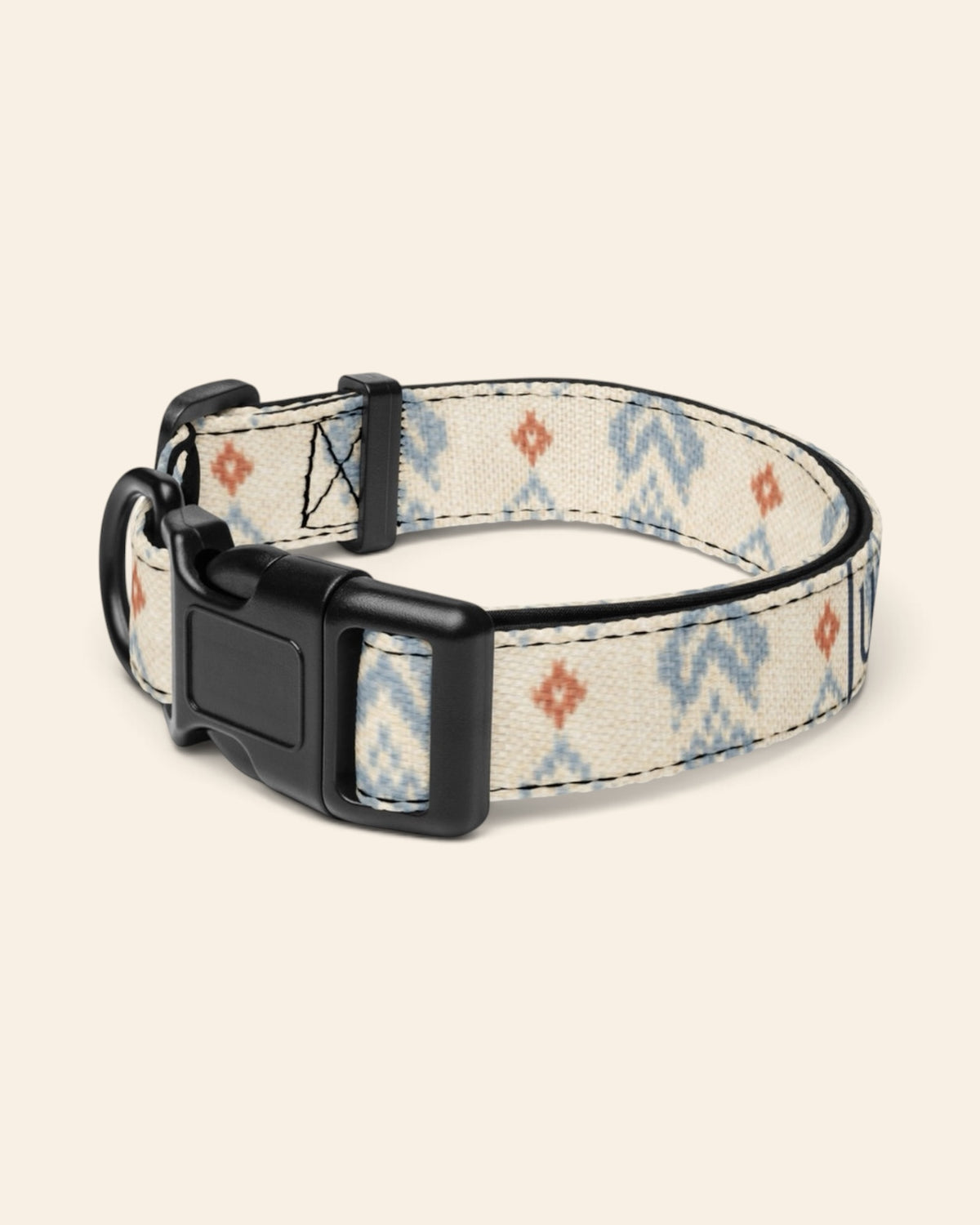 The collar alone on a plain cream background with no personalisation, stood in a ring and angled to show the black side-release buckle face, the D-ring and the tri-glide slider. The webbing is cream, printed with dusty blue zigzag chevrons and stepped diamond motifs with small rust accents.