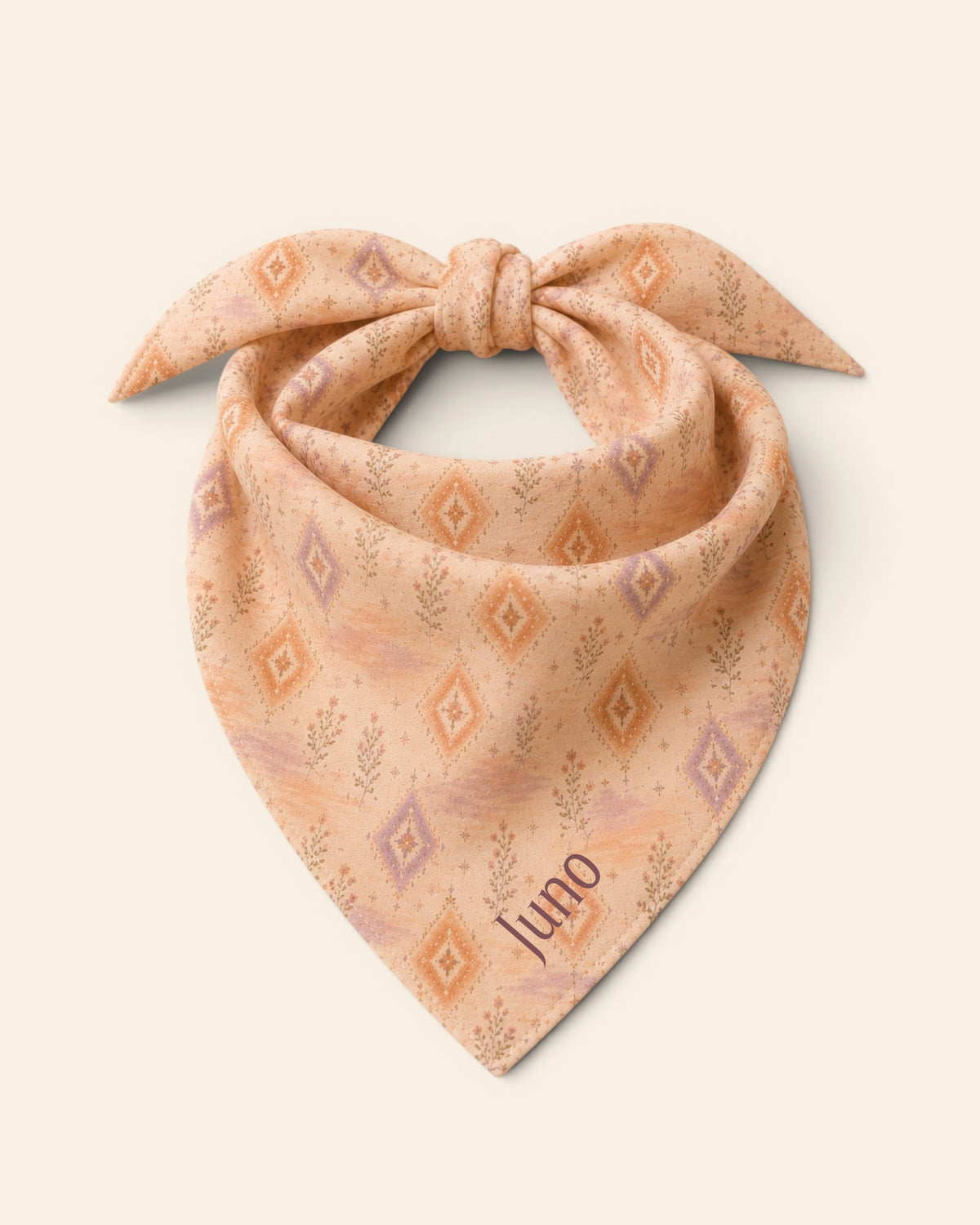 The bandana alone on a plain cream background, knotted so the two tail ends stand up and the triangle hangs below. It is an apricot and peach wash set with cream diamond medallions and fine sprigs, with the sample name Juno printed on the lower right panel.