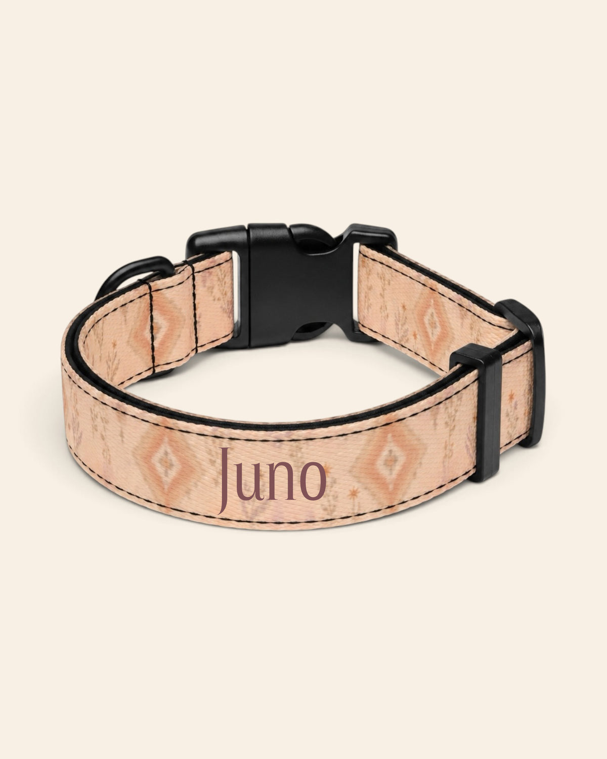 The collar alone on a plain cream background, lying in a loose loop that shows the printed webbing, a black side-release buckle and a black slider. The strap is an apricot and peach wash set with cream diamond medallions and fine sprigs, with the sample name Juno printed along it.