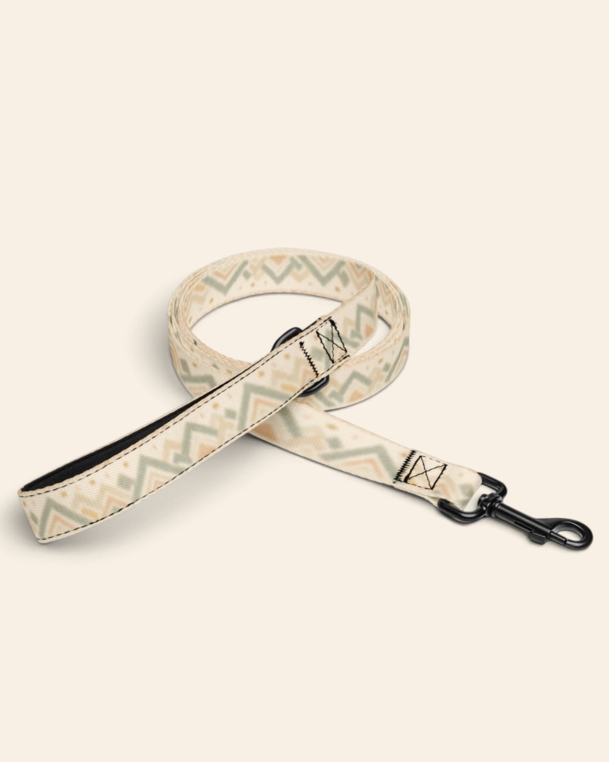The leash coiled on a plain cream background, showing the handle loop, the adjustable slider and the black swivel snap hook. The webbing is cream, banded with sage and apricot chevrons and diamond geometric motifs.