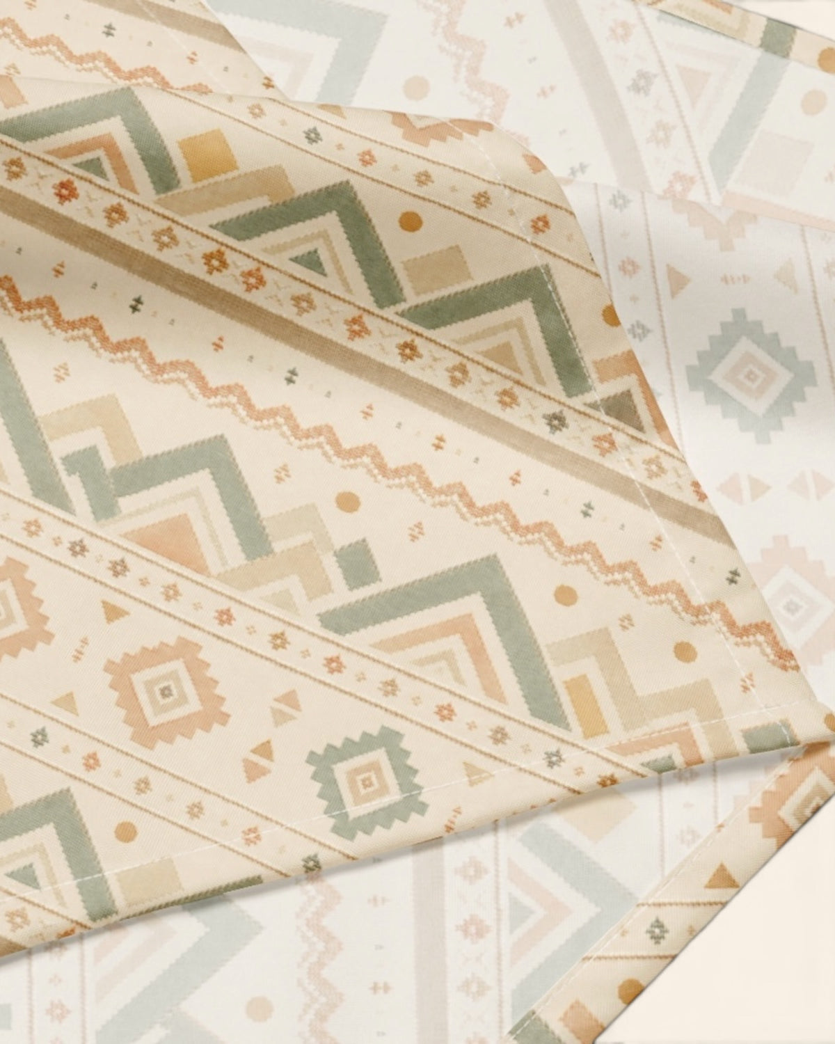 A close crop of the bandana fabric with one corner folded back over a soft cream ground, showing the print at full scale - cream, banded with sage and apricot chevrons and diamond geometric motifs.